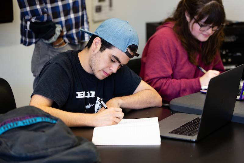 High school student wearing a backwards denim baseball cap and black t-shirt writes in a notebook. A second student wearing glasses and a maroon shirt sits to his left.